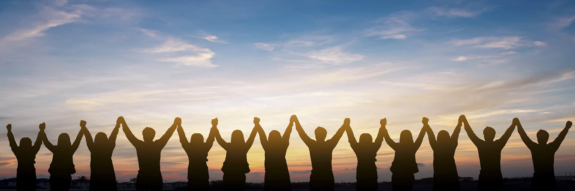 Silhouettes of a group of people standing in a row with their arms raised and hands joined together against a colorful sunset sky.