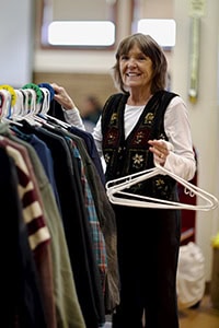 An older woman smiles while sorting clothes on a rack. She holds several empty white hangers in one hand and is surrounded by various jackets and shirts in a well-lit indoor space.