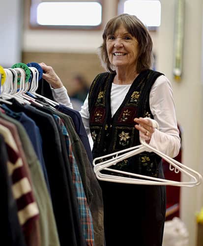 An older woman smiles while sorting clothes on hangers, standing next to a rack of shirts and jackets in a brightly lit room.