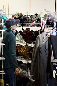 An older person wearing a cap and teal jacket looks at a rack of shoes and clothing in a store, possibly a thrift or secondhand shop.