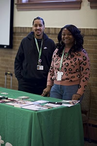 Two people stand behind a green table covered with brochures and pamphlets. Both wear lanyards and smile at the camera; the setting appears to be an indoor event or information fair.