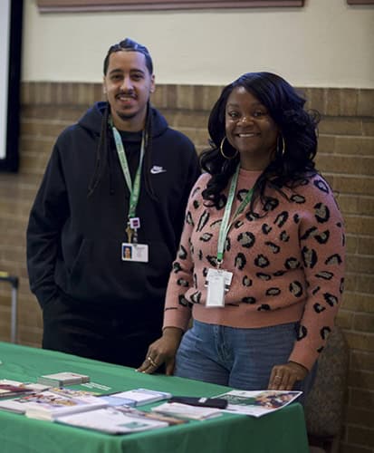 Two people stand behind a green table covered with pamphlets and brochures. Both wear lanyards and smile at the camera; the background features a brick and beige wall.