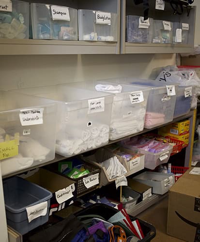 Plastic bins labeled with categories like shampoo, body lotion, and washcloths are neatly organized on shelves, each containing various personal care items and supplies in a storage room.