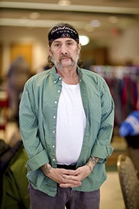 A man with a mustache and tattoos stands indoors, wearing a black bandana, green shirt over a white t-shirt, and dark pants. He clasps his hands in front of him and looks at the camera. Clothing racks are visible in the background.