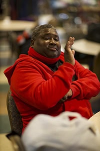 A man wearing a bright red hoodie sits at a table indoors, gesturing with one hand raised near his face. He appears to be engaged in conversation, with a relaxed expression. The background is softly blurred.
