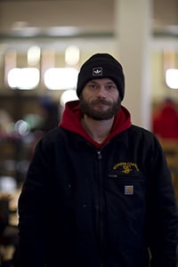 A man with a beard wearing a black beanie, red hoodie, and black jacket stands indoors, looking at the camera with a slight smile. The background is softly blurred.