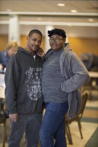 Two people stand close together indoors, smiling at the camera. They are wearing casual clothes and glasses, with one person having an arm around the other. There are tables and people in the background.