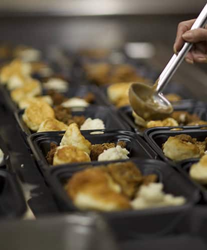A person uses a metal ladle to serve food into black meal trays filled with mashed potatoes and biscuits, lined up on a counter.