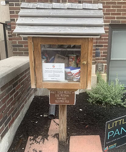 A small wooden food pantry box on a post contains canned goods and packaged food. It stands near a brick building, with a sign below stating it’s an Eagle Scout project completed in December 2021.
