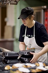 A woman wearing a black cap, glasses, and a white apron stacks black food containers in a kitchen or cafeteria setting, with food trays visible on the counter.
