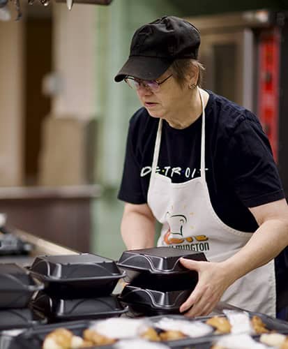 A woman wearing a black cap, glasses, a black shirt, and an apron stacks black takeout containers in a kitchen, with food visible in the foreground.