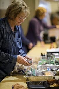 A woman wearing glasses prepares food at a table with aluminum trays and packaged items, while another person works in the background.