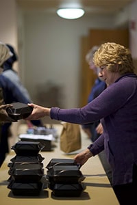 A woman wearing a purple sweater hands a food container to someone across a table, with several stacked black food containers in front of her. Other people are visible in the background, helping with distribution.