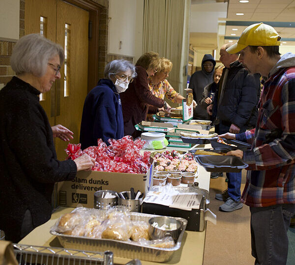 Volunteers serve food to people standing in line at an indoor community event. Various dishes and bread are arranged on tables, and people are receiving meals and interacting with the servers.