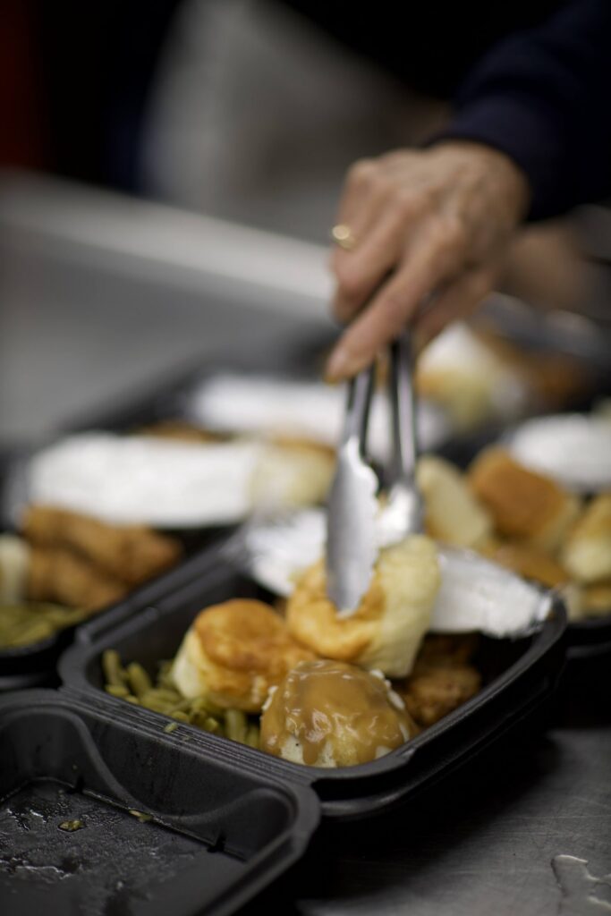 A volunteer’s hands serving a hot lunch into a take-out container at Outreach on 7th